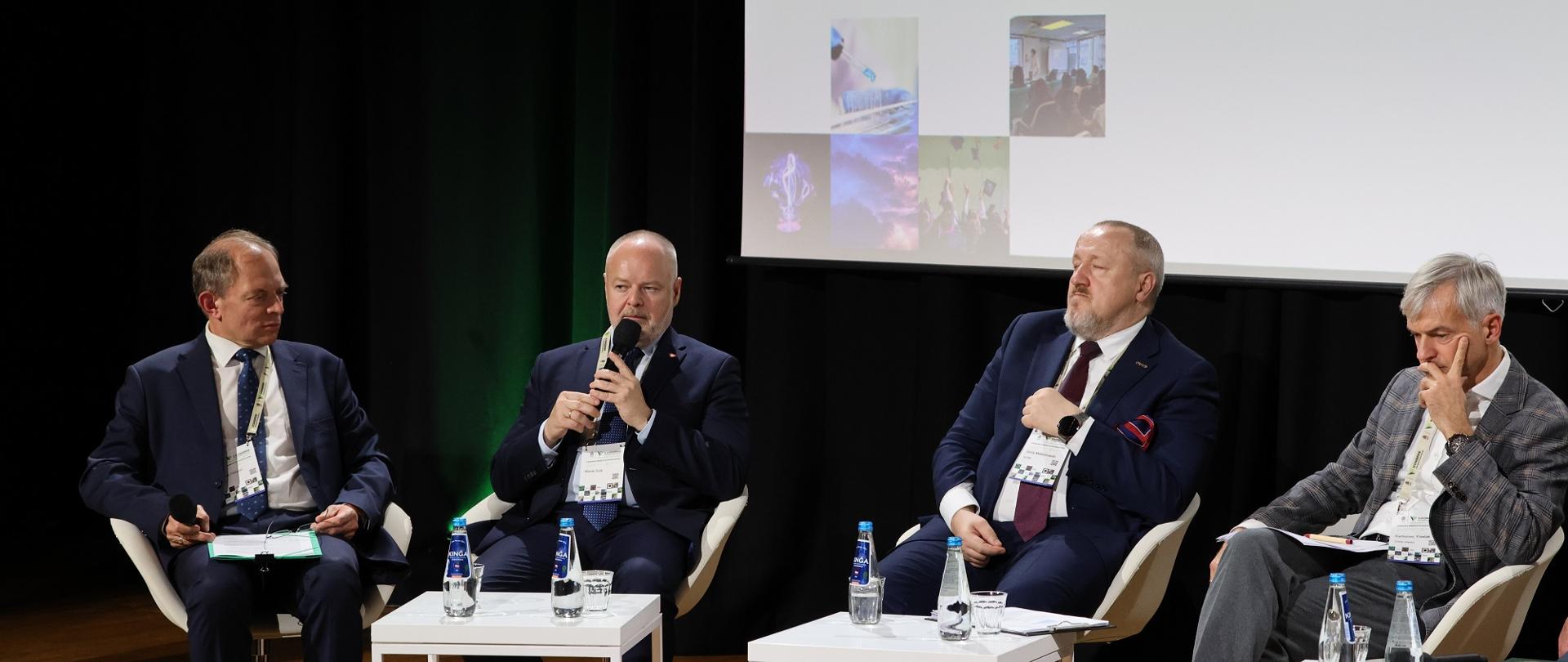 Na sali za małymi białymi stolikami siedzi na fotelach czterech mężczyzn w garniturach, drugi od lewej wiceminister Gzik mówi do trzymanego w ręku mikrofonu.