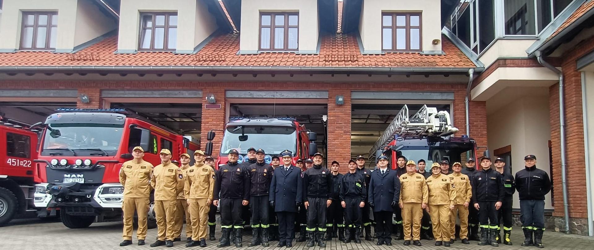 Zdjęcie przedstawia zmiany służbowe oraz pozostałych funkcjonariuszy i pracowników cywilnych Komendy Powiatowej Państwowej Straży Pożarnej w Gołdapi, zlokalizowanych na placu zewnętrznym komendy po zmianie służby z okazji przejścia na zaopatrzenie emerytalne st. ogn. Grzegorza Franke. W tle widać pojazdy pożarnicze zlokalizowane w garażu Komendy Powiatowej Państwowej Straży Pożarnej w Gołdapi