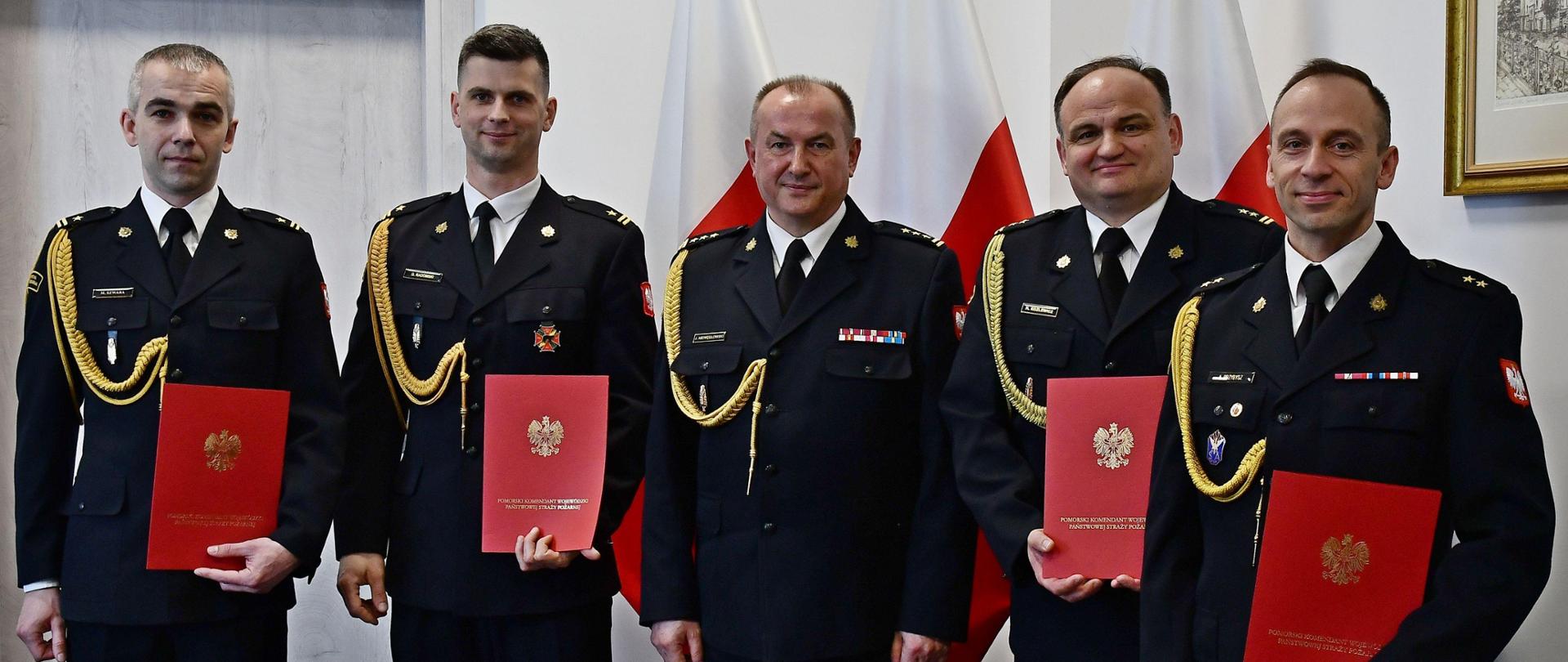 Dwóch strażaków w mundurach wyjściowych ze sznurem stoi obok siebie mężczyzna w środku jest bez teczki za nimi ustawione są trzy flagi Polski wisi obraz oraz znajdują się drzwi. 