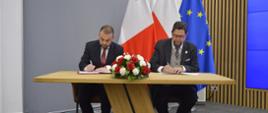 Za drewnianym stolikiem siedzą wiceminister Szeptycki i mężczyzna w czarnym garniturze, podpisują otwarte dokumenty, przed nimi na stoliku bukiet biało-czerwonych kwiatów, za nimi pod szarą ścianą flagi Polski i UE.