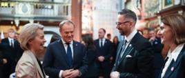 W dużej sali z wieloma obrazami na ścianach stoi minister Kulasek, rozmawia z kobietą w beżowej marynarce, obok nich stoi premier Tusk, dookoła dużo elegancko ubranych osób.