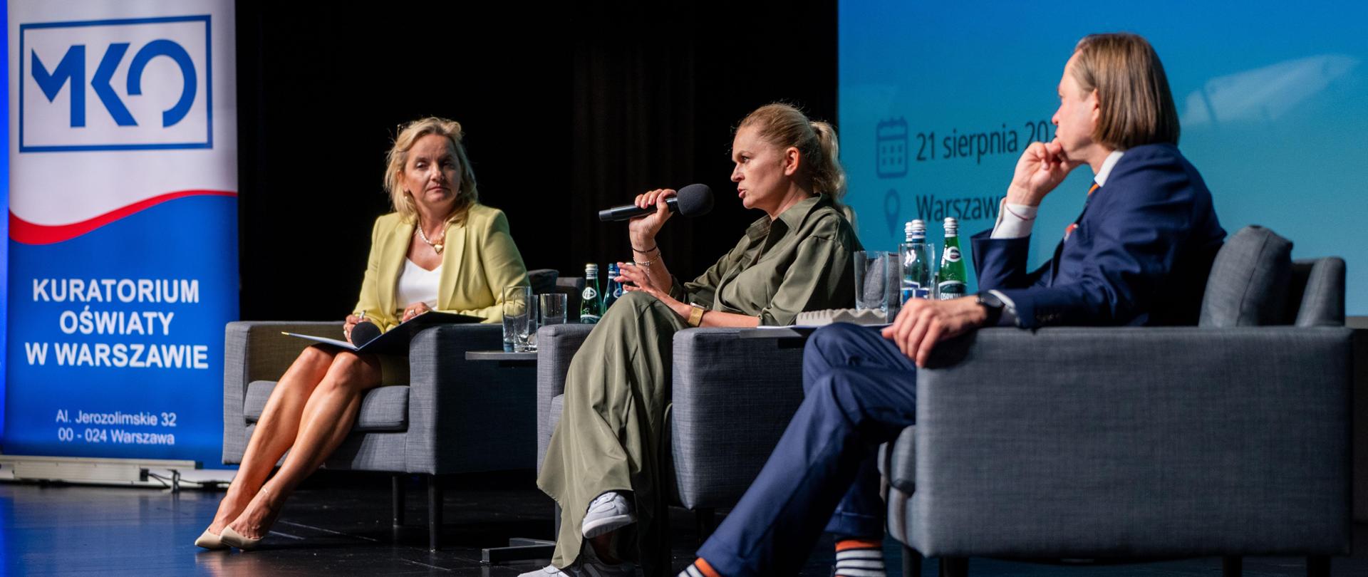 Na scenie ustawione są trzy fotele. Pośrodku siedzi minister Barbara Nowacka i przemawia do mikrofonu. Po jej lewej stronie siedzi mężczyzna a po prawej kobieta, którzy przysłuchują się wystąpieniu i patrzą na minister edukacji. W tle widać logo Kuratorium Oświaty w Warszawie. 