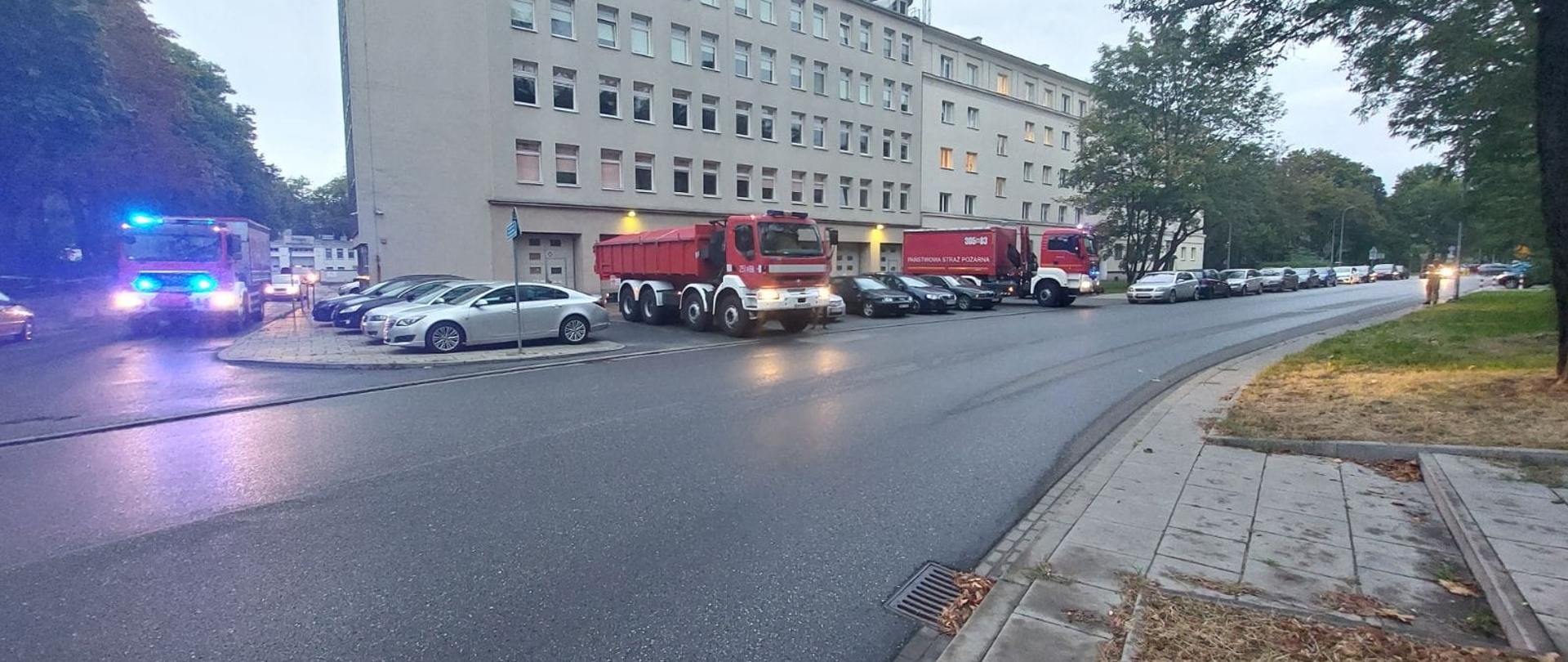  Zdjęcie wykonane na otwartej przestrzeni. W tle ściana żółtego budynku o wysokości 5 kondygnacji. Przed budynkiem stoją licznie zaparkowane cywilne samochody. Pomiędzy nimi czerwone samochody ciężarowe. Samochody znajdują się w ruchu i mają na dachach zapalone niebieskie migające sygnały świetlne.
