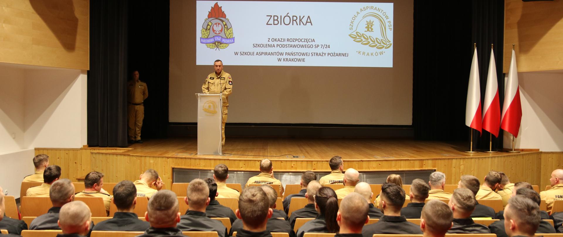 Aula SA PSP w Krakowie. Uczestnicy zbiórki z okazji rozpoczęcia szkolenia podstawowego w zawodzie strażak SP 7/24. Strażacy widziani od tyłu. Na scenie nadbryg. Piotr Filipek w czasie okolicznościowego przemówienia. Na slajdzie tytułowym informacja o charakterze i powodzie zbiórki. 