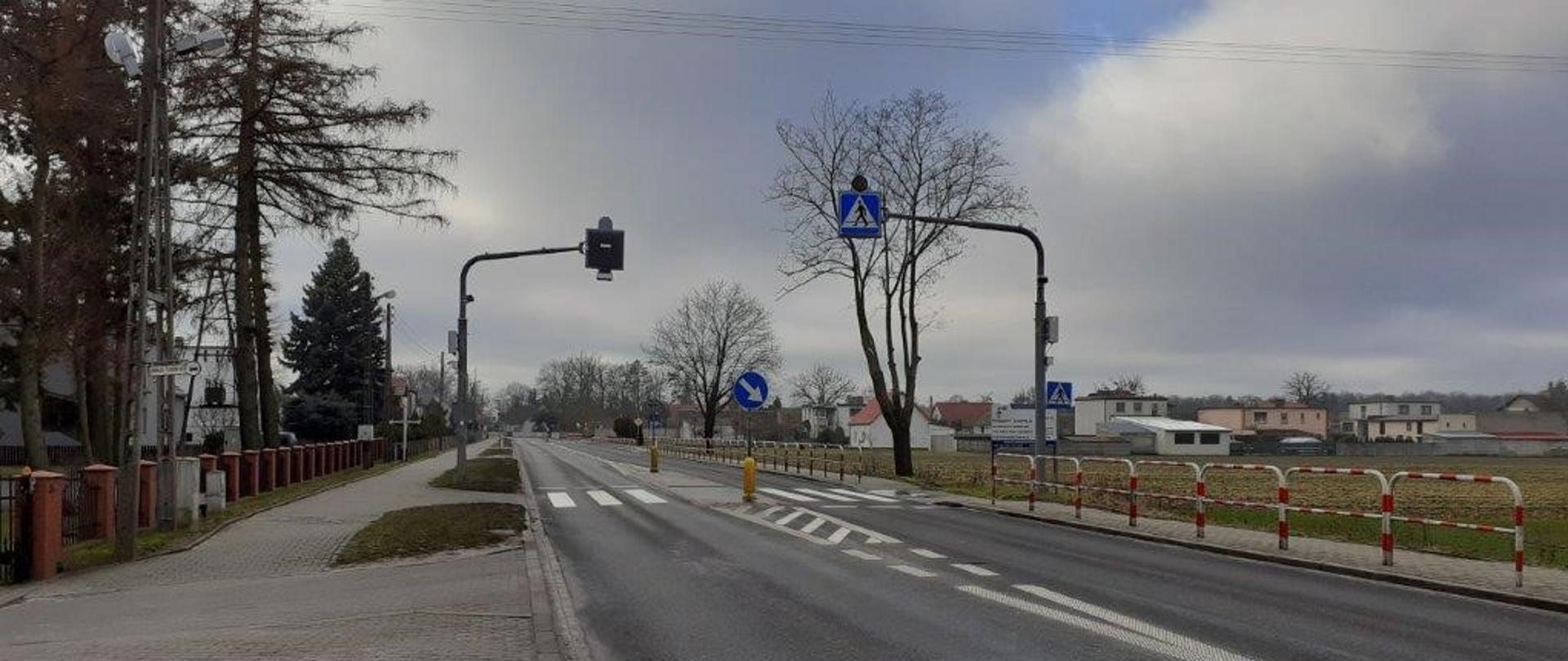 DK92 Bolewice. Z prawej strony widoczna droga oraz barierki prowadzące do przejścia dla pieszych. Znaki widoczne na wysięgniku nad drogą.