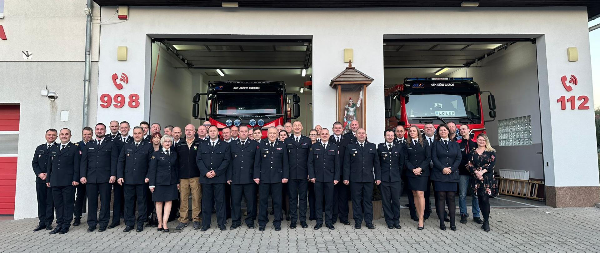 Strażacy ubrani w mundury wyjściowe stoją na zdjęciu grupowym przed remizą ochotniczej straży pożarnej