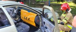 Zdjęcie przedstawia samochód osobowy z otwartymi drzwiami pasażera. Deska rozdzielcza zabezpieczona jest przed wystrzałem poduszki powietrznej specjalną kurtyną w kolorze żółtym. Strażak w ubraniu specjalnym i czerwonym hełmie mocuje kurtynę do nadkola pojazdu.