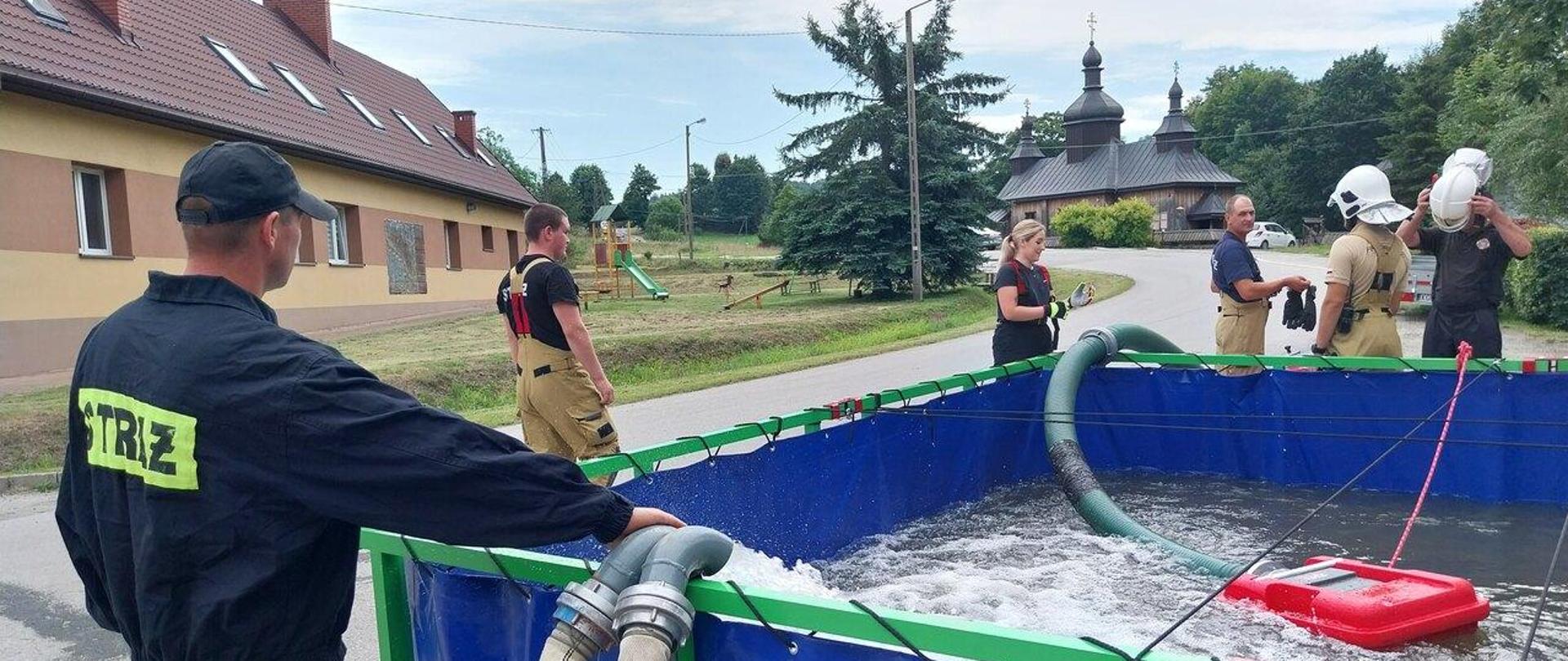 Na zdjęciu widoczni strażacy ubrani w ubrania koloru czarnego i piaskowego przy zbiorniku wodnym w trakcie jego napełniania podczas ćwiczeń pożarniczych w zakresie dostarczania wody na duże odległości w miejscowości Bartne. Zbiornik na wodę koloru niebieskiego w tle widoczna cerkiew i budynek ochotniczej Straży Pożarnej w Bartnem.