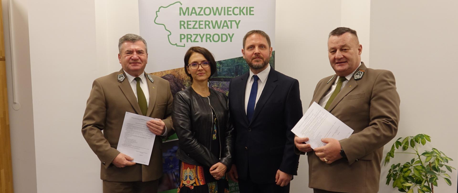 Trzech mężczyzn i kobieta stojących na tle roll-upa z logo Mazowieckich rezerwatów przyrody