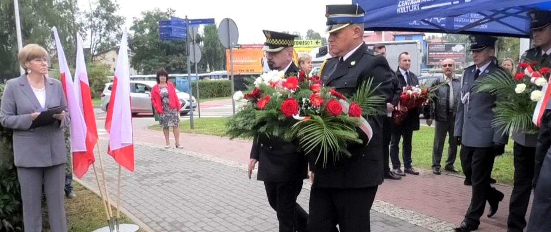 Strażak i strażnik miejski stoją i trzymają w rękach kwiaty. Za nimi stoją policjanci . w tle widać kobietę i dwóch mężczyzn. Na trawniku po lewej stroni stoi kobieta . Obok niej są trzy biało-czerwone flagi.