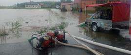 Miejscowośc Pakówka. Na pierwszym planie dwie pompy szlamowe, ktorymi odpompowywana jest woda z zalanego pola. Z prawej strony stoi przyczepka z pompa o wysokiej wydajności. W tle budynki, a przy nich ludzie, ciągnik rolniczy i auto dostawcze. Widoczny jest też wał ułozony z worków wypełnionych piaskiem. 