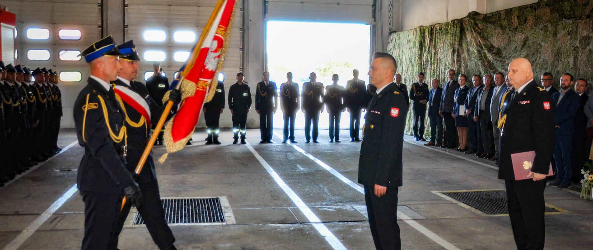 młodszy brygadier Konrad Krupa całuje sztandar komendy za nim stoi starszy brygadier Piotr Rudecki, na przeciwko niego stoi poczet sztandarowy, w tle strażacy, zdjęcie wykonane w garażu jednostki ratowniczo-gaśniczej