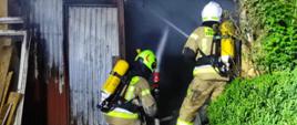 Na zdjęciu widocznych jest dwóch strażaków wyposażonych w aparaty ochrony układu oddechowego. Strażacy podają prądy wody do wnętrza budynku gospodarczego. 