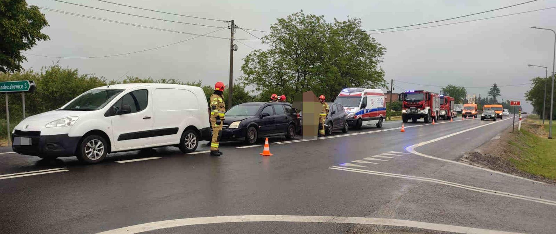 Zdjęcie zrobione w dzień na drodze. Na zdjęciu widać rozbite samochody, strażaków , karetkę pogotowia ratunkowego, samochody strażackie. Pada deszcz. 