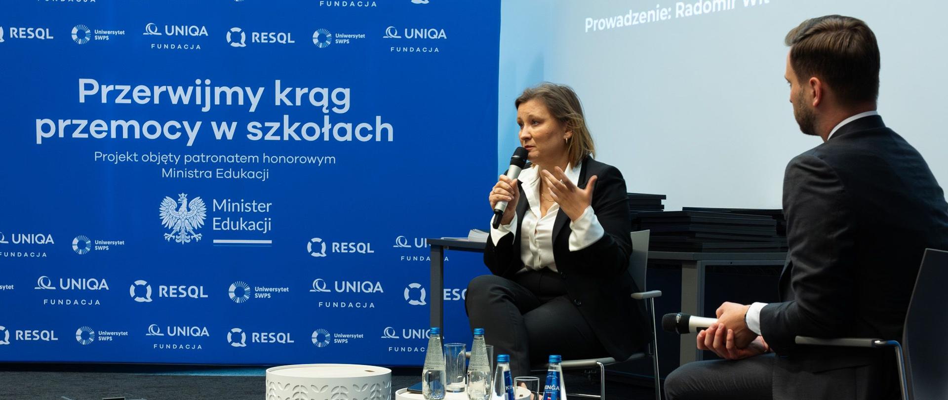 Wiceminister Paulina Piechna-Więckiewicz przemawia na siedząco do mikrofonu. Obok niej siedzi dziennikarz prowadzący panel dyskusyjny. W tle widać niebieską ściankę z napisem "Przerwijmy krąg przemocy w szkołach", z logo organizatorów, a także z informacją o patronacie honorowym MEN. 