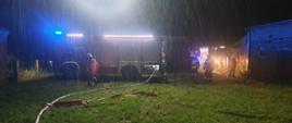 Zdjęcie przedstawia dwa samochody ratowniczo-gaśnicze, które oświetlają teren działań oraz węże gaśnicze podłączone do samochodu stojącego z lewej strony zdjęcia. W pobliżu samochodów widać czterech strażaków. Pada deszcz. Pora nocna.