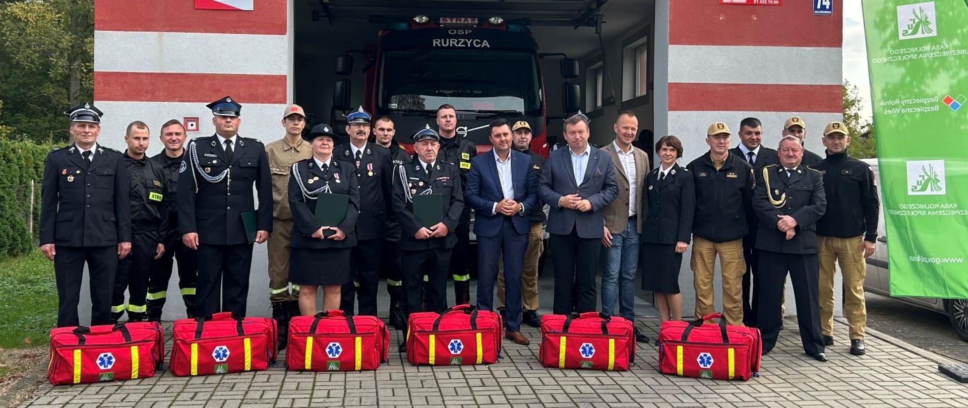 Duża grupa osób, w tym większość w mundurach galowych i strojach koszarowych straży pożarnej pozuje na tle garażu i stojącego w nim wozu bojowego. Obok rollup z logo KRUS, a przed nimi sześć toreb z zestawami ratownictwa przedlekarskiego.