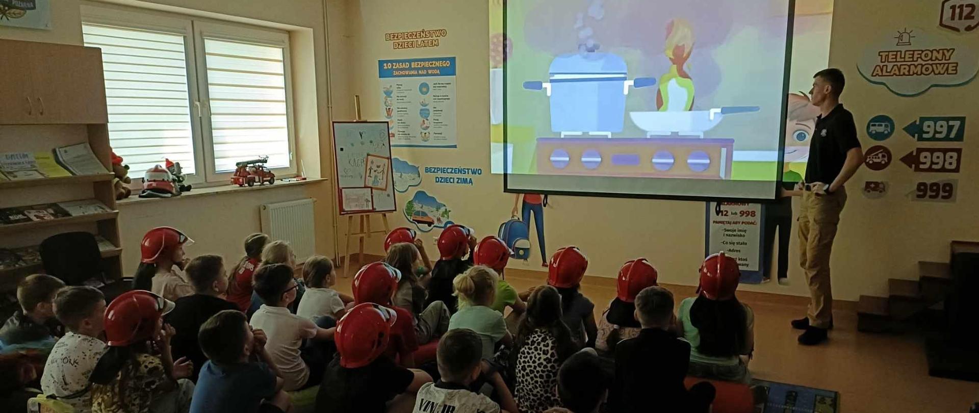 Zdjęcie przedstawia dzieci podczas zajęć edukacyjnych w sali OGNIK. Dzieci obserwują wyświetlaną prezentację. Na slajdzie pokazany jest pożar tłuszczu na patelni. Jeden ze strażaków tłumaczy dzieciom zasady zachowania podczas tego typu pożarów.