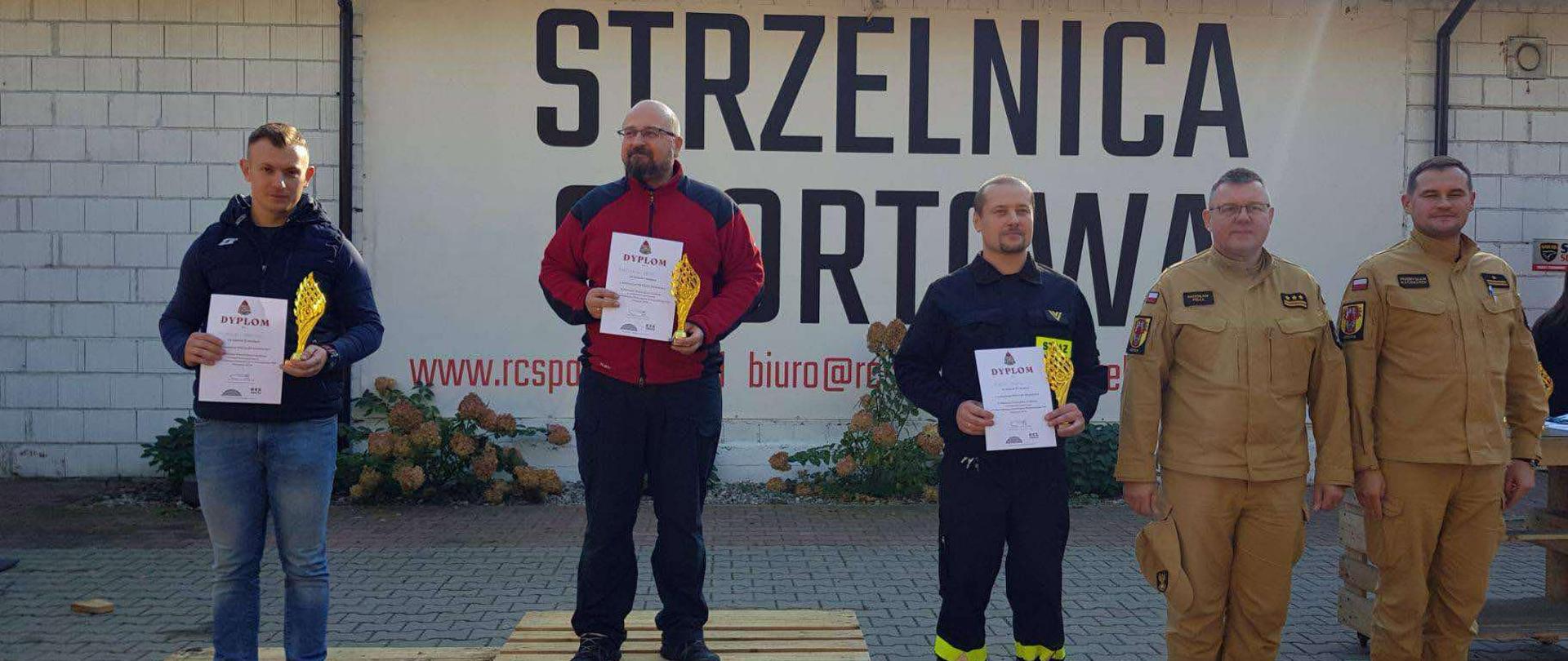 trzech zawodników, którzy wygrali w swojej konkurencji stojących na podium z palet, obok stoi dwóch komendantów