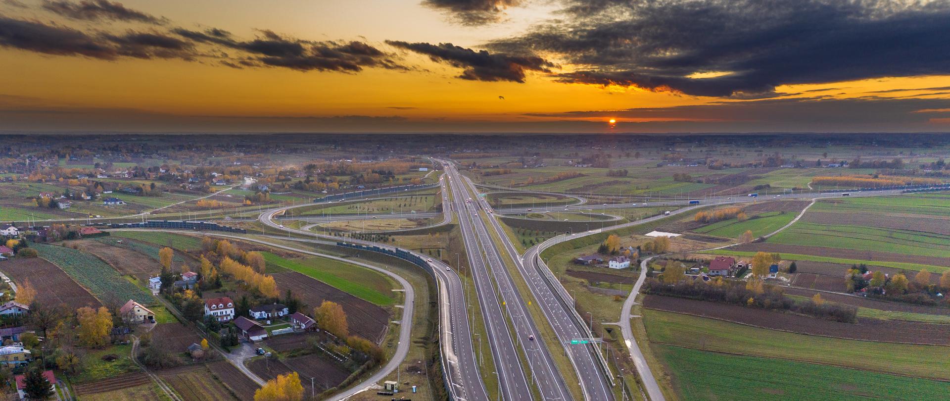 Zdjęcie lotnicze węzła Lublin Sławinek na połączeniu dróg ekspresowych S12 i S17. Zdjęcie wykonane przy zachodzie Słońca. Widoczny ruch pojazdów na obu drogach ekspresowych. W tle widoczne zabudowania oraz pola uprawne i łąki.