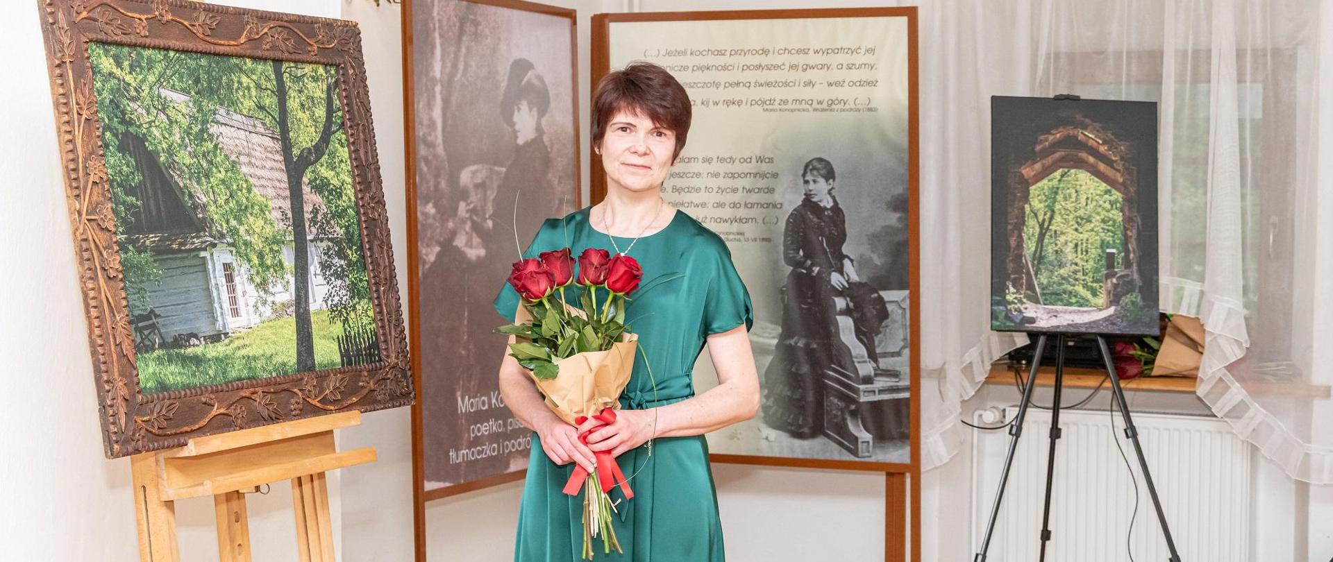 Na zdjęciu stoi słuchaczka Studium w zielonej sukience. W rękach trzyma czerwone róże. W tle obrazy i zdjęcia.