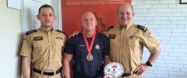 Zdjęcie przedstawia trzech strażaków na tle baneru, jeden z nich ma w ręku i na szyi medale z wygranych zawodów.