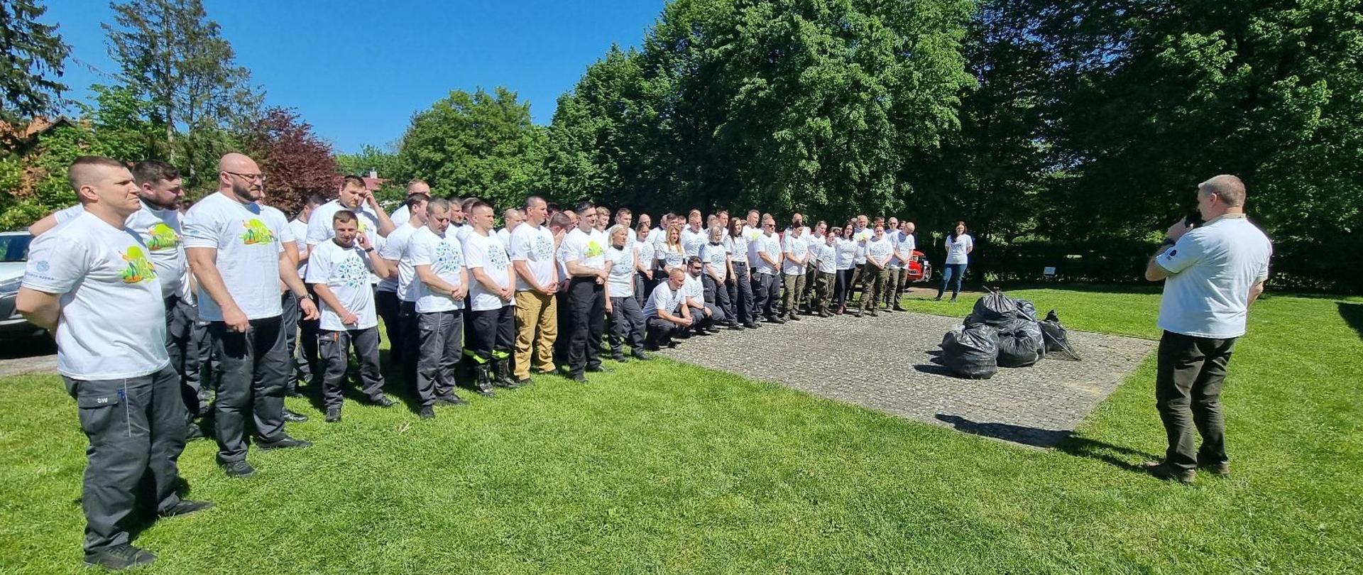 Prawie sto osób w białych koszulkach stoi na trawie, przed nimi mężczyzna tłumaczący zasady Operacji czysta rzeka. W tle drzewa i błękitne niebo