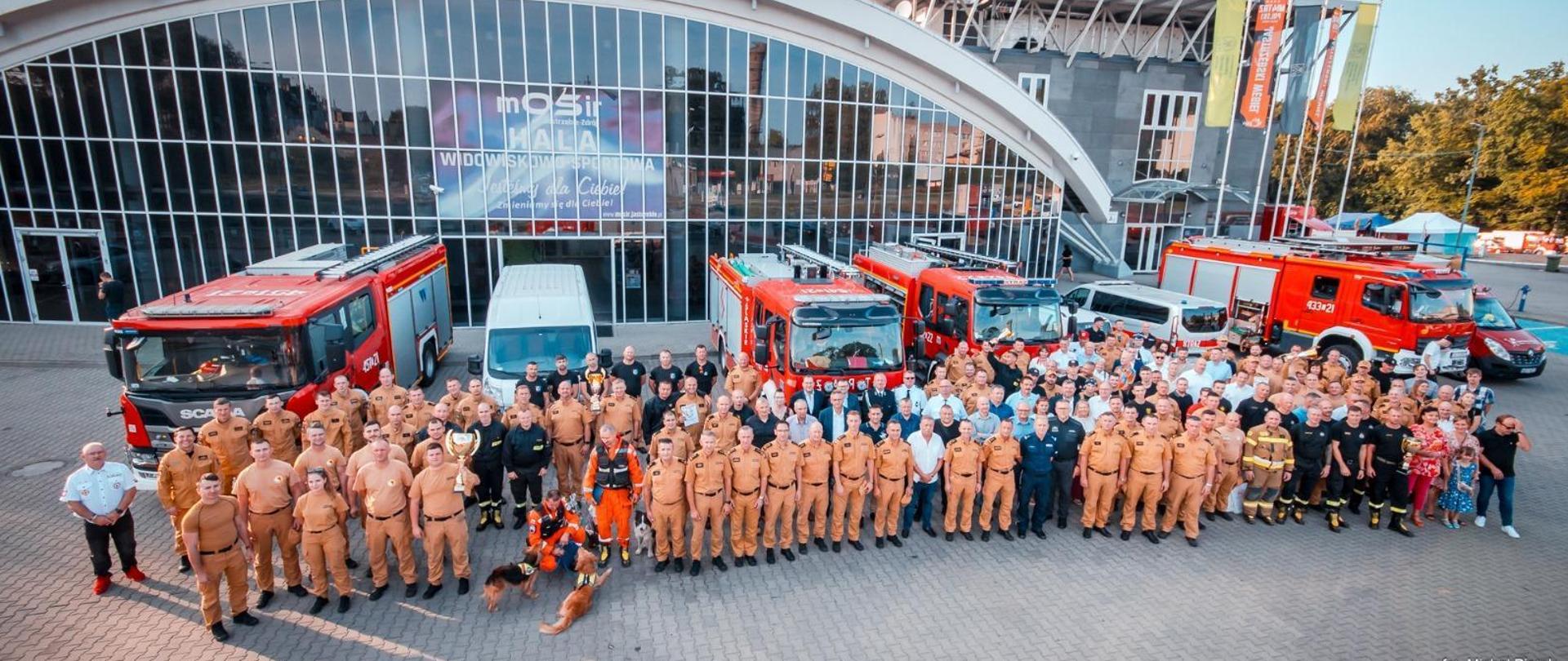 Zdjęcie przedstawia zgromadzonych uczestników zawodów w Ratownictwie Technicznym. W tle samochody pożarnicze i hala widowiskowo - sportowa.