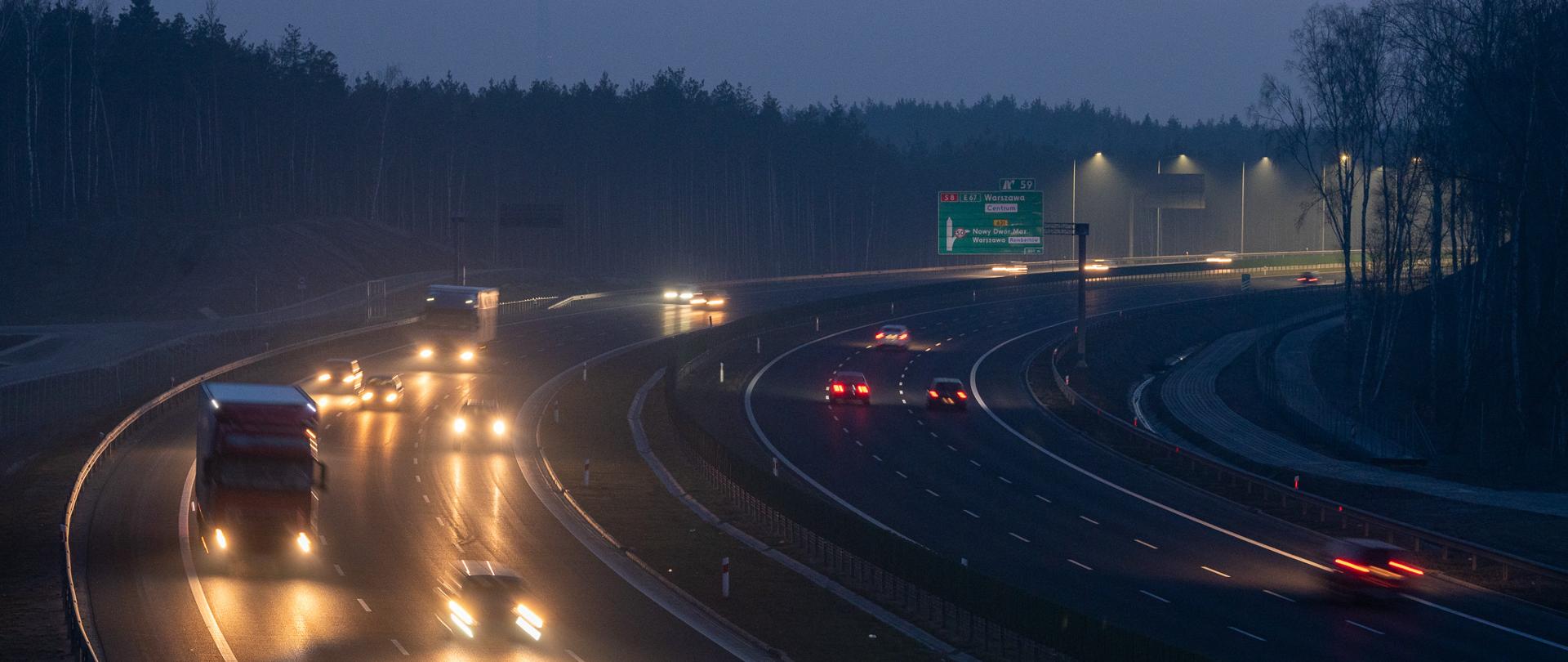 Zdjęcie przedstawia drogę ekspresową o zmierzchu, oświetloną światłami samochodów i latarni ulicznych. Na drodze widoczne są różne pojazdy, w tym ciężarówki i samochody osobowe, jadące w obu kierunkach. W tle widać las, a po prawej stronie drogi znajduje się zielony znak drogowy z informacjami o kierunkach jazdy.