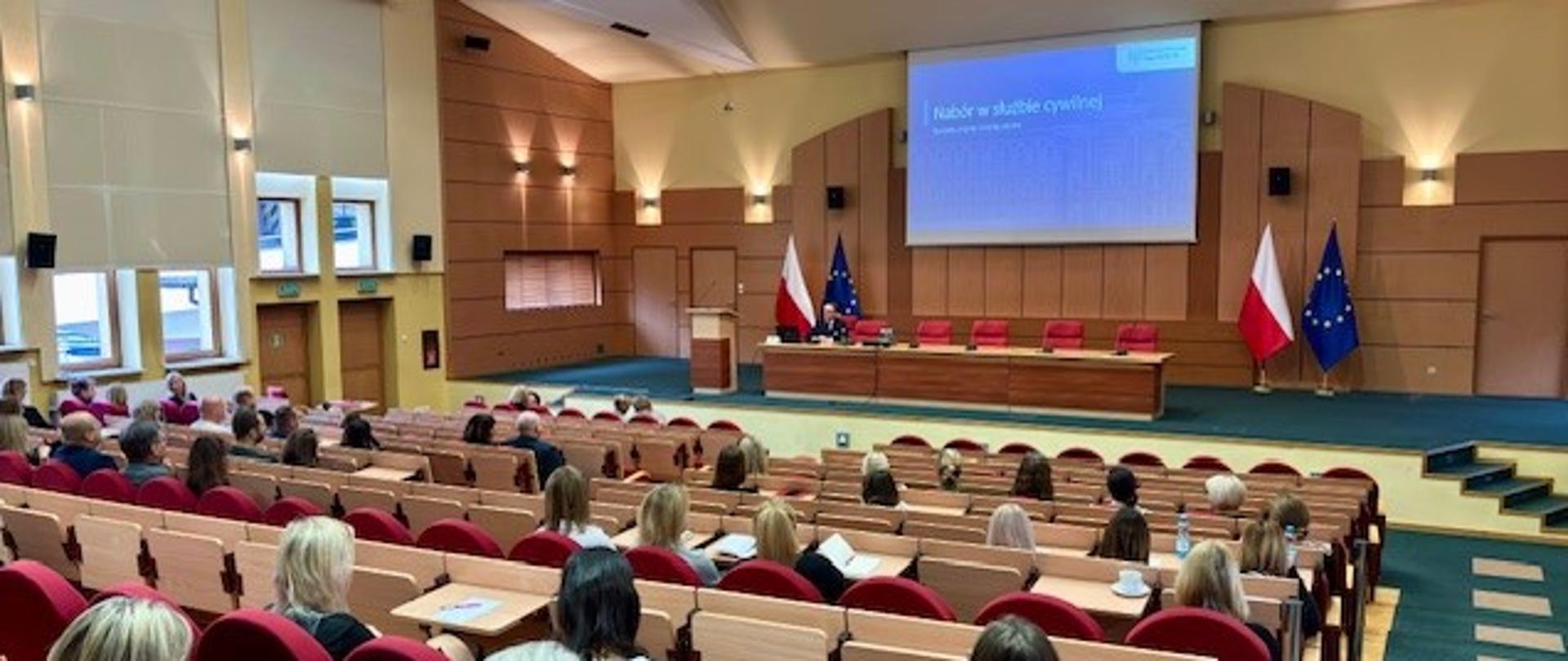 Sala wykładowa. Na pierwszym planie słuchacze siedzą w rzędach. Na drugim planie stół konferencyjny na podium. Przy stole siedzi mężczyzna. Za nim widać prezentacje multimedialna na dużym ekranie.