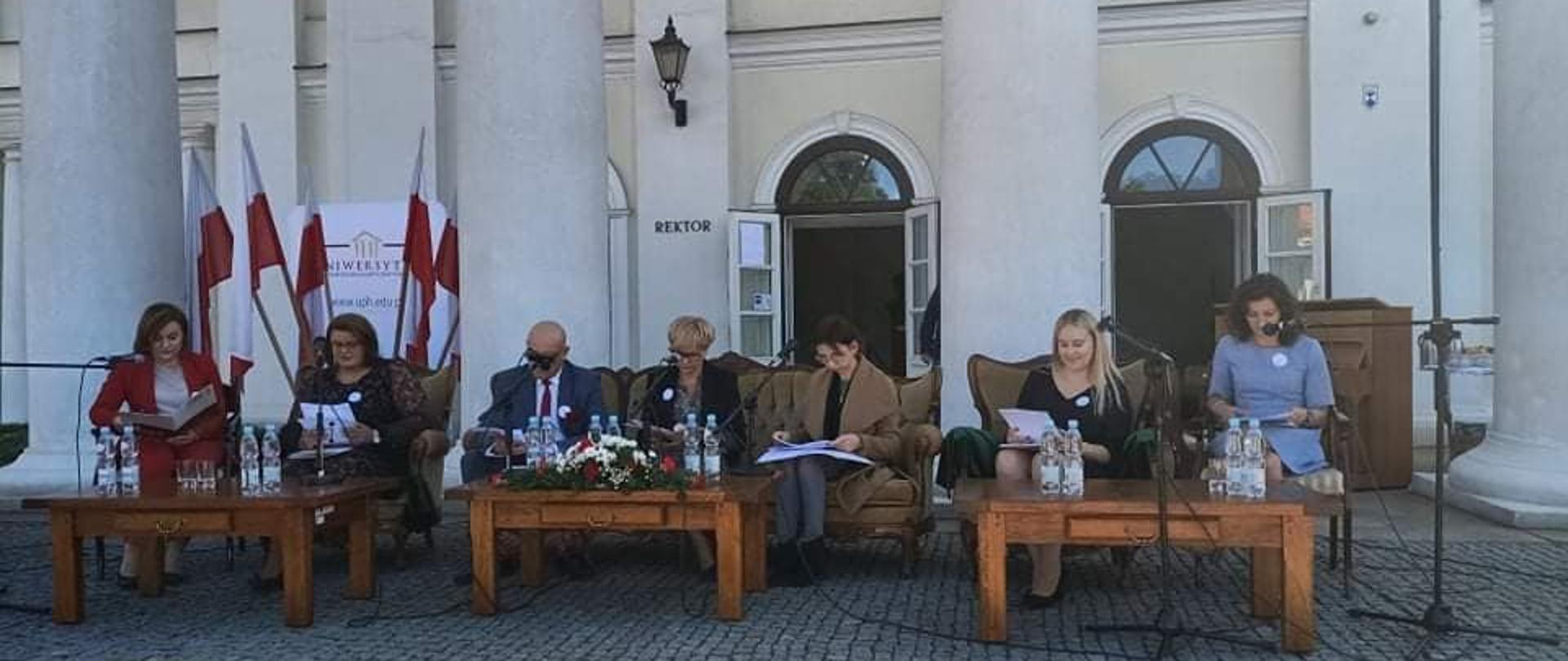 Na zdjęciu znajduje się siedem osób. Siedzą oni za trzema stolikami, trzymając w rękach białe kartki. W tle znajduje się fragment frontowej części gmachu Pałacu Ogińskich w Siedlcach z kolumnami. 