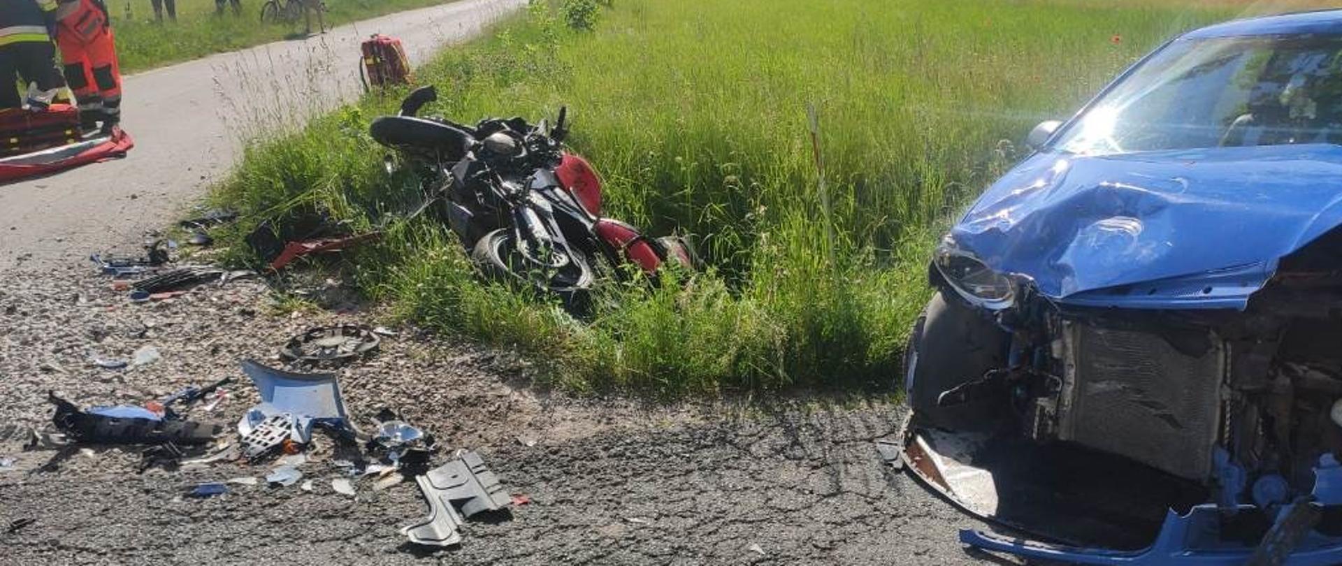 Na zdjęciu uszkodzony motocykl w rowie, rozbity samochód na poboczu drogi, na drugim planie strażacy wraz z ZRM udzielają pomocy medycznej poszkodowanemu motocykliście