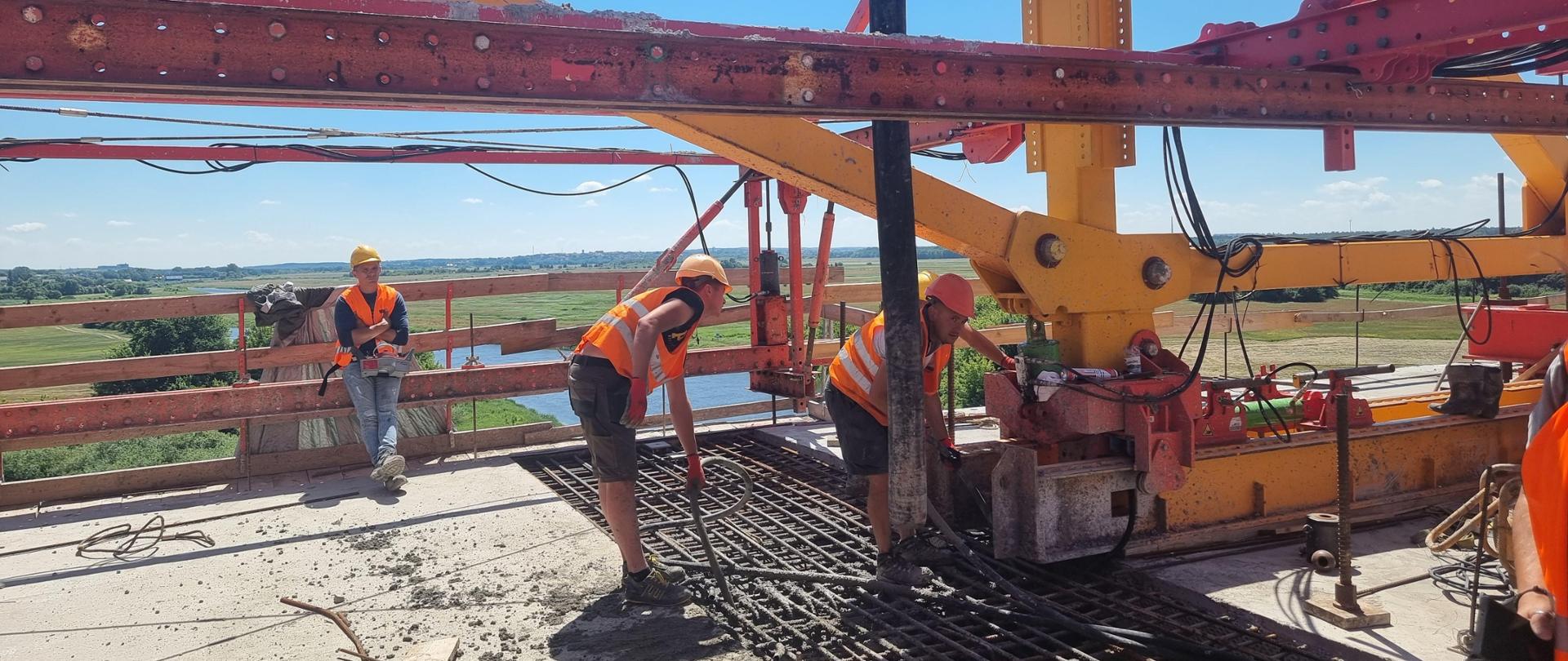 pracownicy budowlani w kaskach i pomarańczowych kamizelkach betonują zazbrojone połączenie mostu nad Narwią. Pracownik dystybuuje beton przy pomocy pompy do betonu, drugi zagęszcza beton zagęszczarką buławową