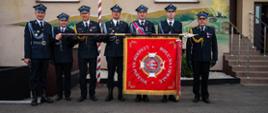 Na zdjęciu widzimy grupę siedmiu druhów w mundurach stojących przed budynkiem jednostki. Trzymają między sobą duży sztandar, na którym widnieje haftowany tekst i symbole, w tym hełm strażacki i dwa skrzyżowane topory. Sztandar jest głównie czerwony z złotymi i białymi akcentami. W tle widać fasadę budynku z oknami oraz fragment muralu przedstawiającego zieleń.