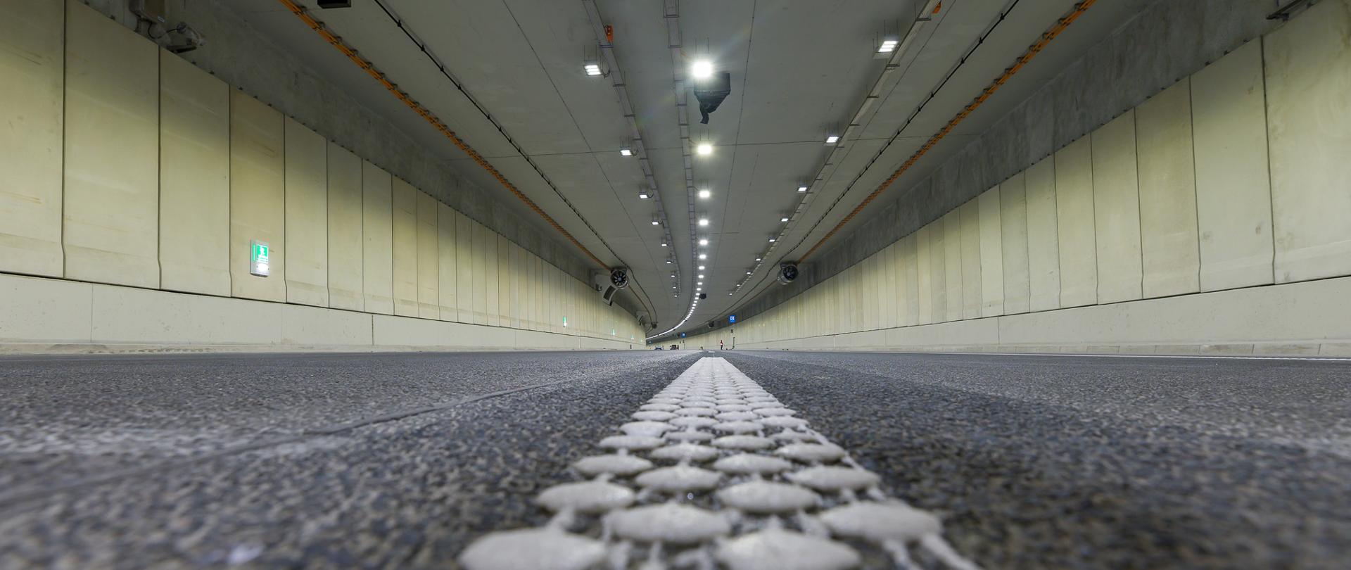 Ujęcie na wnętrze tunelu pod Ursynowem. Widoczne ściany tunelu z zamontowanymi urządzeniami technicznymi i oświetleniem.