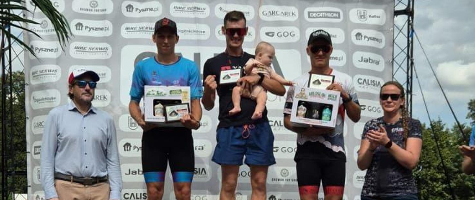 Trzech mężczyzn w strojach sportowych stoi na podium, mężczyzna w środku trzyma na ręku niemowlę, po lewej stronie podium stoi mężczyzna w czapce z daszkiem i ciemnych okularach, po prawej stronie podium stoi kobieta klaszcząc w dłonie