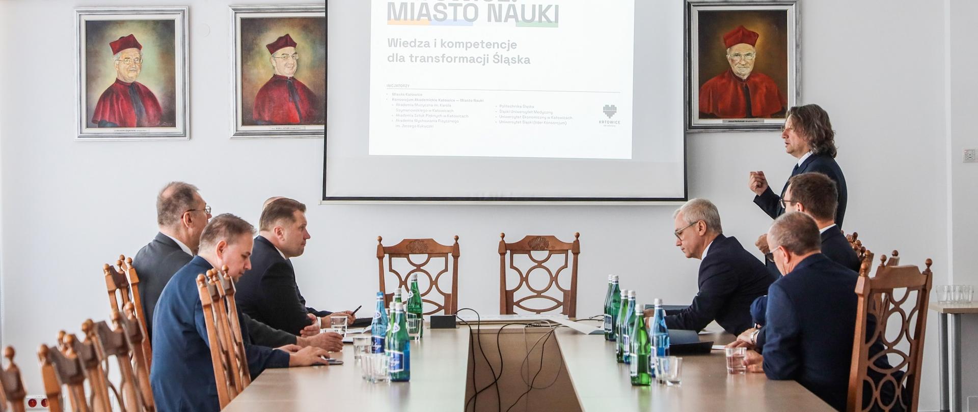 Po obu stronach drewnianego stołu siedzi grupa ludzi, obok stołu ekran z napisem Katowice miasto nauki.