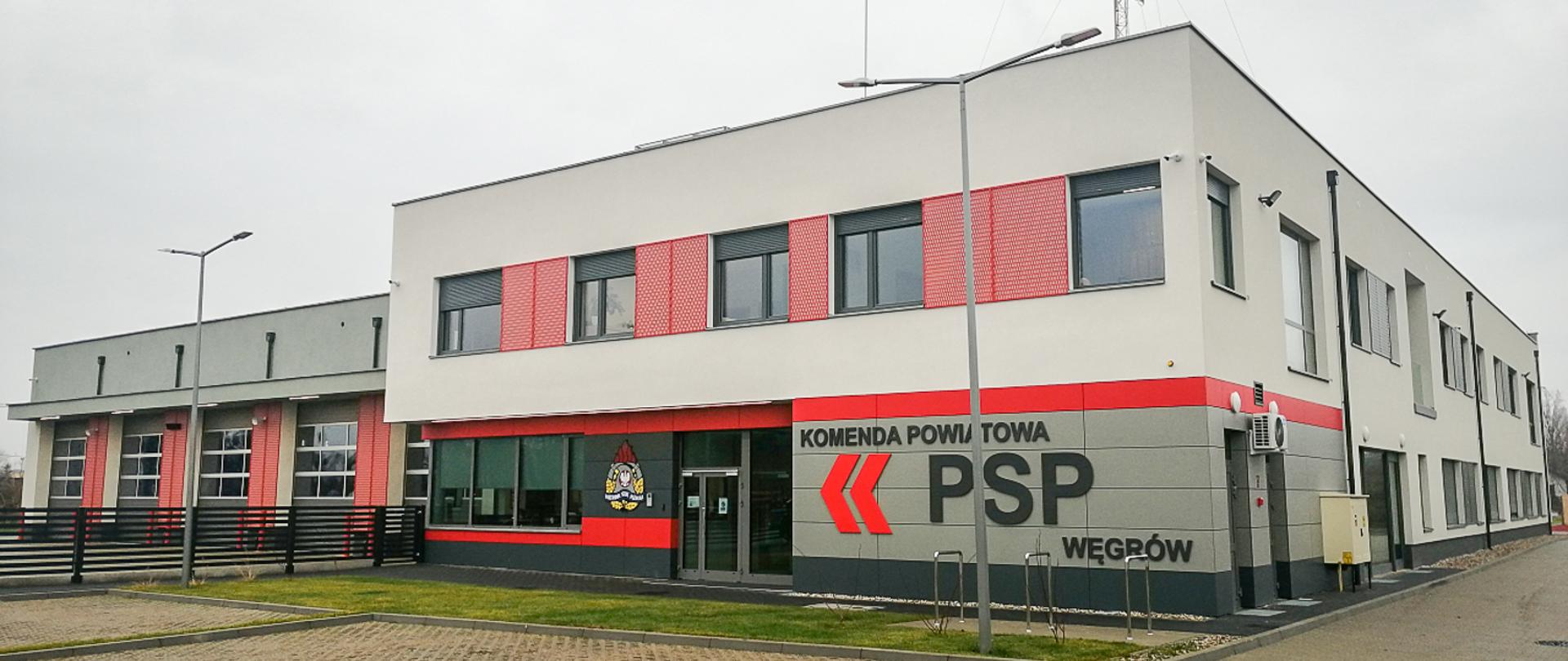 Zdjęcie przedstawia budynek Komendy Powiatowej PSP w Węgrowie w kolorystyce szaro-czerwonej. Na pierwszym planie wejście do obiektu. Pop prawej stronie wejścia napis Komenda Powiatowa PSP Węgrów. Po lewej stronie wejścia logotyp Państwowej Straży Pożarnej.