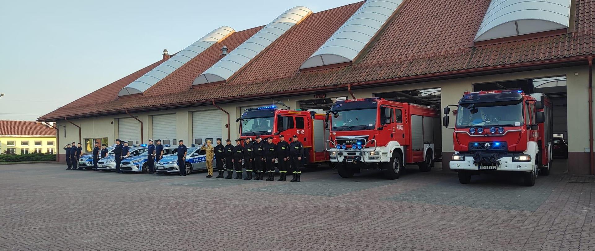 Strażacy i policjanci stojący przed samochodami i radiowozami w rzędzie obok siebie. W tle garaż.