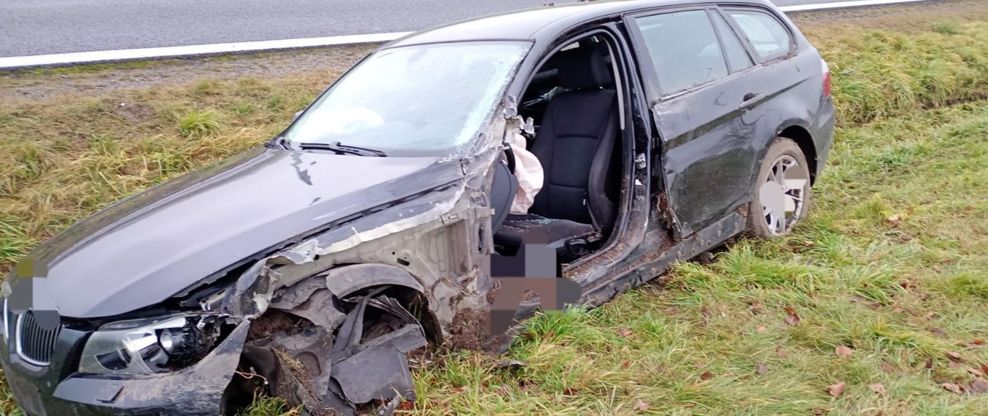 Na zdjęciu ciemny samochód osobowy, który stoi w rowie. Ma uszkodzony lewy przód oraz nie ma drzwi od strony kierowcy. W tle widać jezdnie asfaltową. Jest widno.