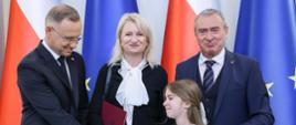Prezydent Duda podaje rękę małej dziewczynce w białej koszulce, obok niego stoi kobieta w czarnej bluzce i mężczyzna w granatowym garniturze, za nimi flagi Polski i UE.