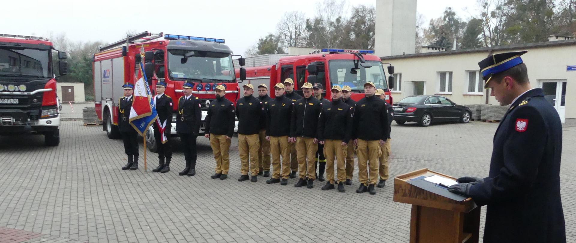 Na zdjęciu Zastępca Komendanta Powiatowego odczytuje list okolicznościowy z Komendy Głównej dotyczący obchodów Narodowego Święta Niepodległości. Na drugim planie stoi poczet sztandarowy oraz funkcjonariusze jednostki ratowniczo-gaśniczej. W oddali widać pojazdy Komendy. 