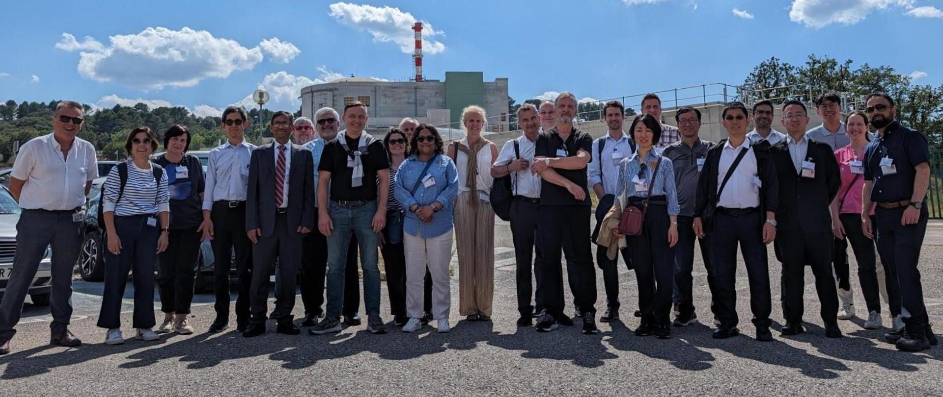 Uczestnicy 51. Posiedzenia Komitetu ds. Działalności Dozorów Jądrowych stoją w grupie podczas wizyty studyjnej na terenie reaktora RJH (Jules Horowitz Reactor) w Cadarache. W tle budynek reaktora oraz biało-czerwony komin