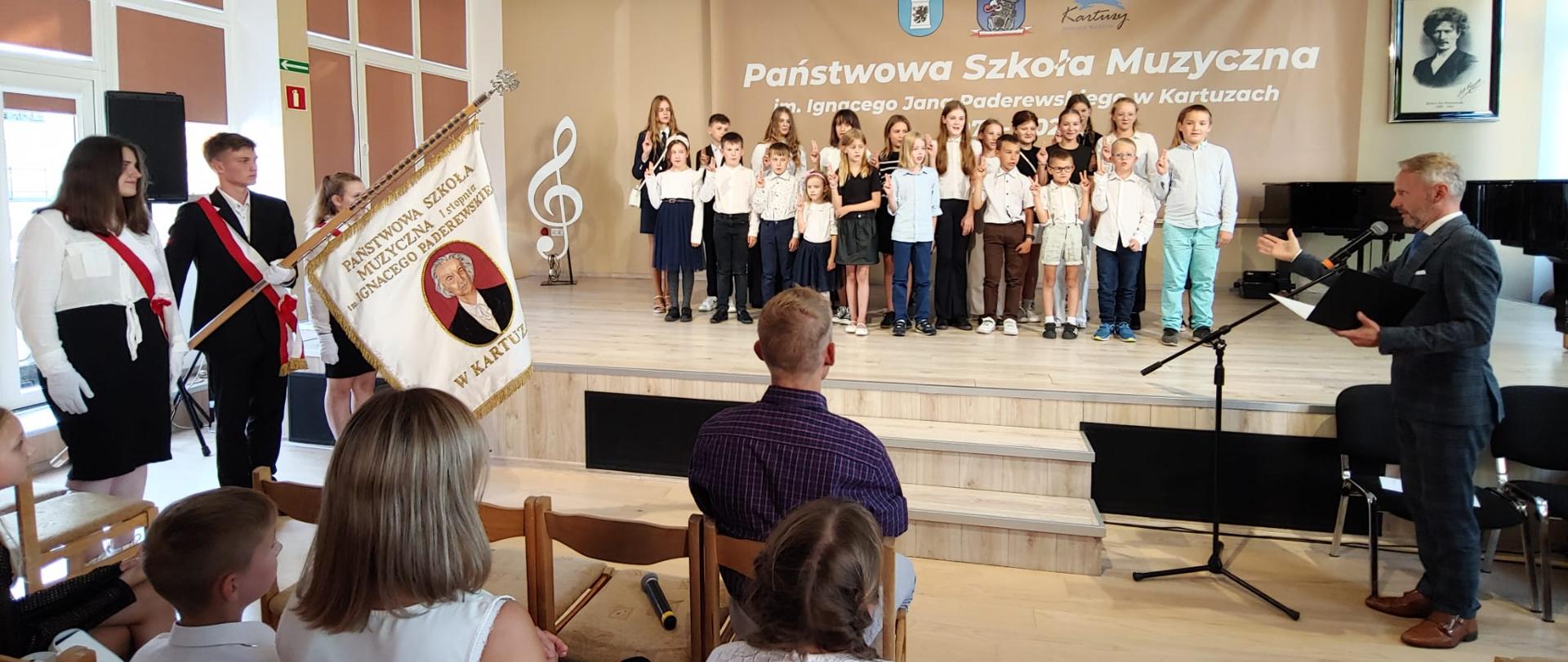 na scenie uczniowie klas pierwszych, po lewej stronie poczet sztandarowy, po prawej dyrektor szkoły