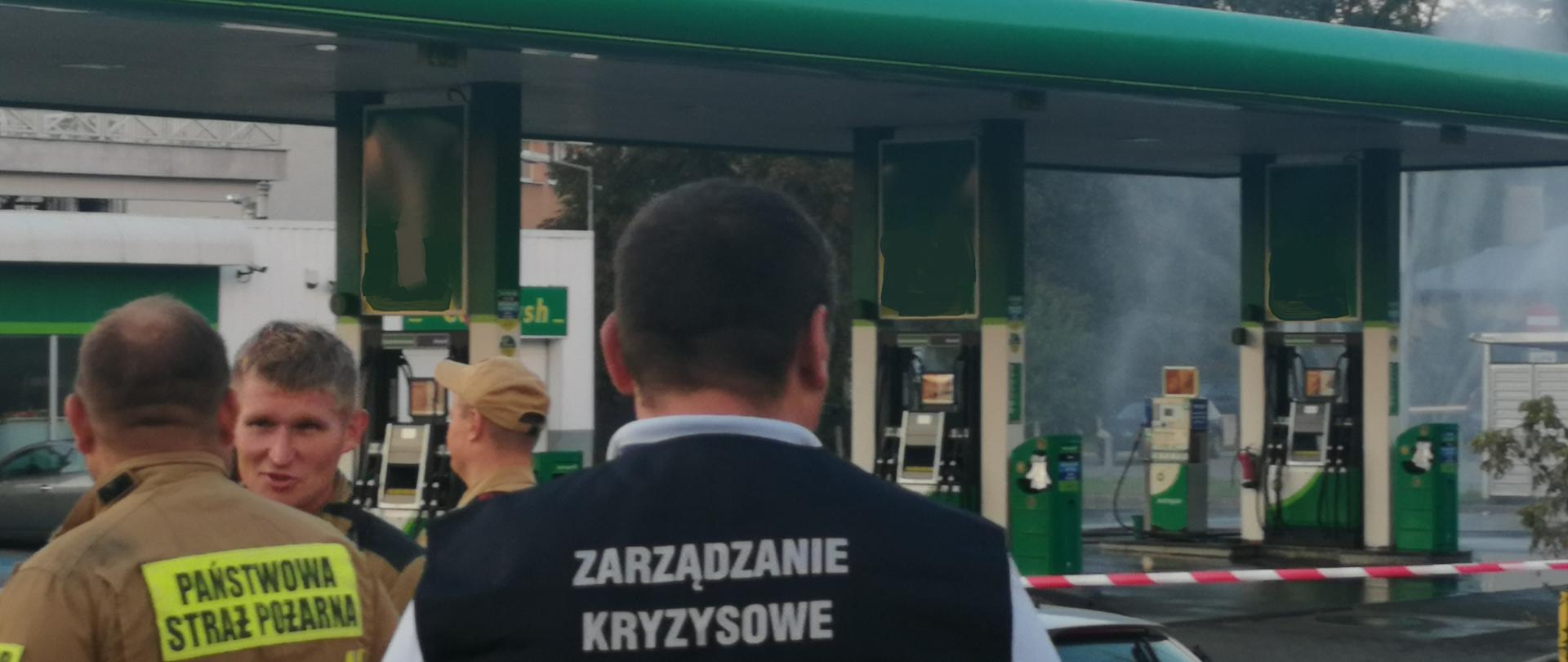 Pracownik zarządzania kryzysowego oraz trzej Funkcjonariusze Państwowej Straży Pożarnej monitorują miejsce wycieku LPG. W tle widać stację benzynową a za nią wysoką na kilka metrów kurtynę wodną.