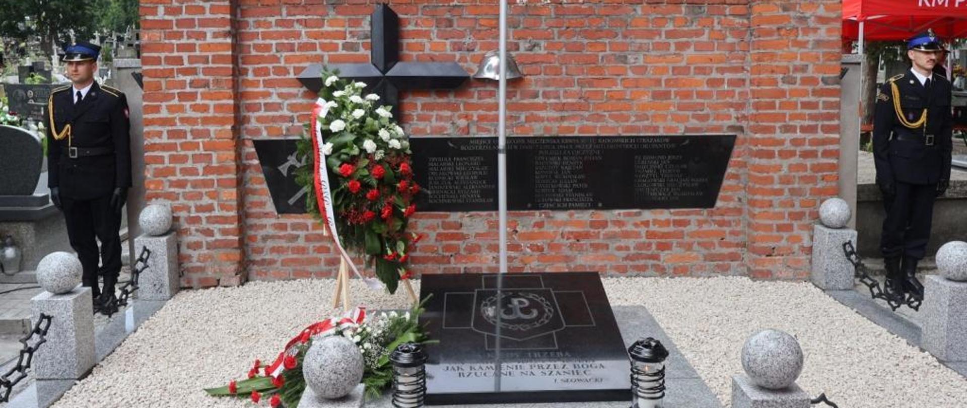 Zdjęcie wykonane na cmentarzu komunalnym. Na zdjęciu widać pomnik zamordowanych przez hitlerowskich żołnierzy strażaków. Na płycie położony jest wieniec w kolorze zielonym z biało – czerwoną szarfą. Po prawej i po lewej stronie pomnika stoi strażak ubrany w mundur galowy w kolorze ciemnym granatowym. Pełnią wartę honorową podczas uroczystości. 