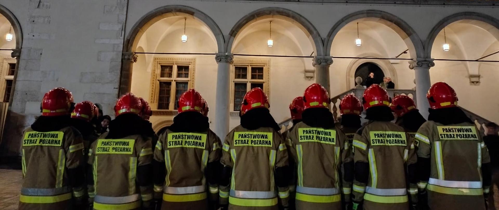 Dziedziniec Zamku Królewskiego na Wawelu – 14 stojących tyłem do autora zdjęć strażaków – osób ubranych w piaskowego koloru ubrania specjalne z czerwonymi hełmami na głowach. 