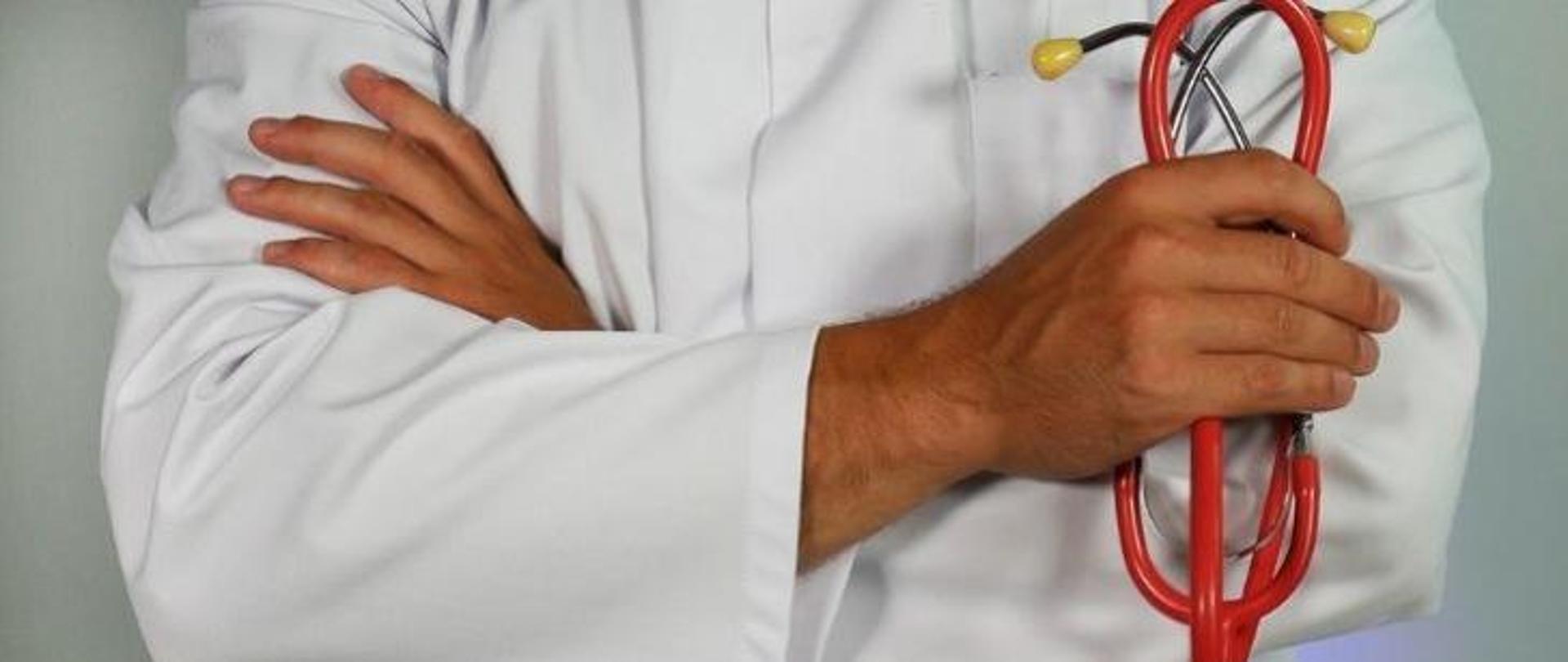 Sylwetka lekarza, najprawdopodobniej mężczyzny, który jest ubrany w biały fartuch medyczny, a w ręce trzyma czerwony stetoskop 