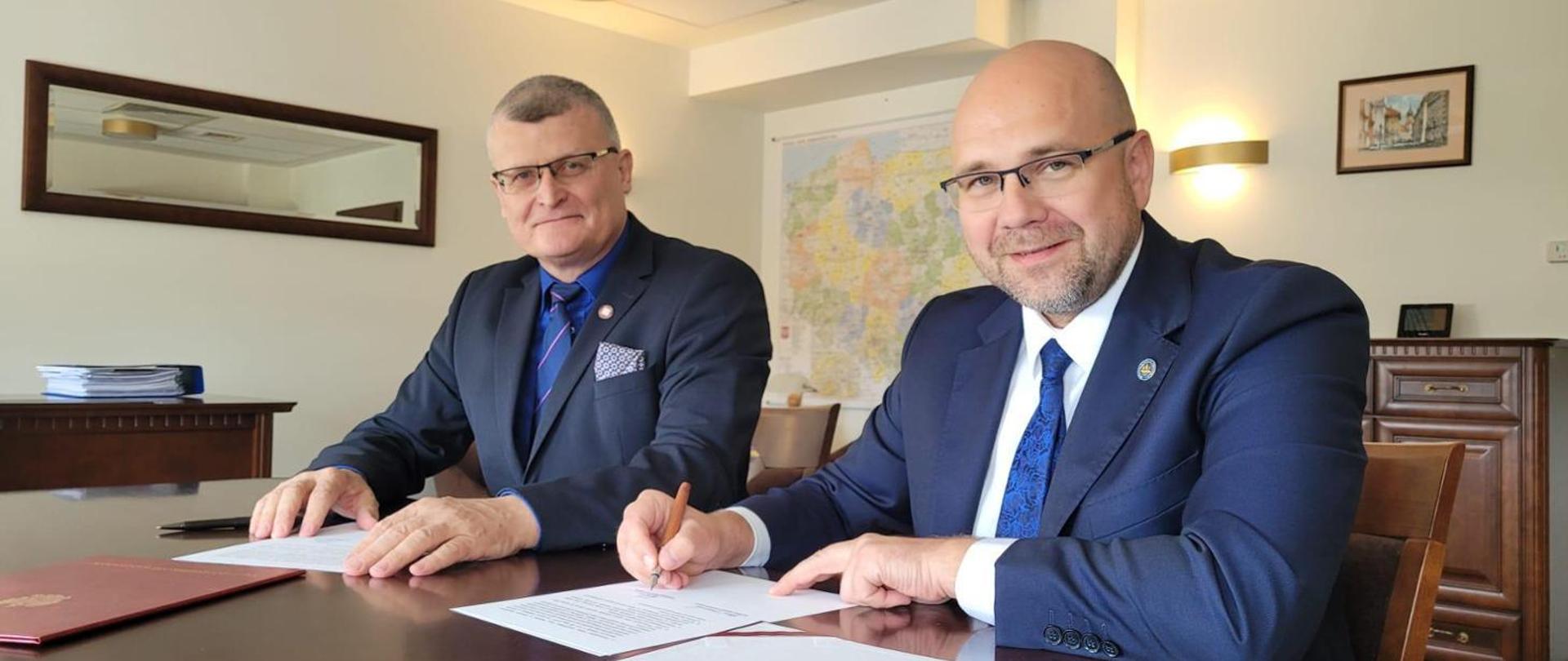 dwóch uśmiechniętych mężczyzn siedzących za biurkiem podpisujących dokument
