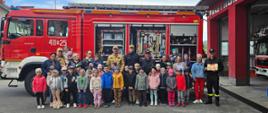 Kolorowa fotografia wykonana na zewnątrz w pogodny dzień. Przedstawia grupę przedszkolaków które odwiedziły strażnicę PSP oraz strażaków w ubraniach służbowych na tle samochodu pożarniczego.